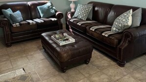 A living room with two brown leather sofas, a patterned rug, a window with a view of trees, and a floral painting on the wall.