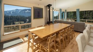 Una sala moderna con una gran mesa de comedor de madera, un sofá gris y vistas a un paisaje montañoso a través de amplios ventanales.