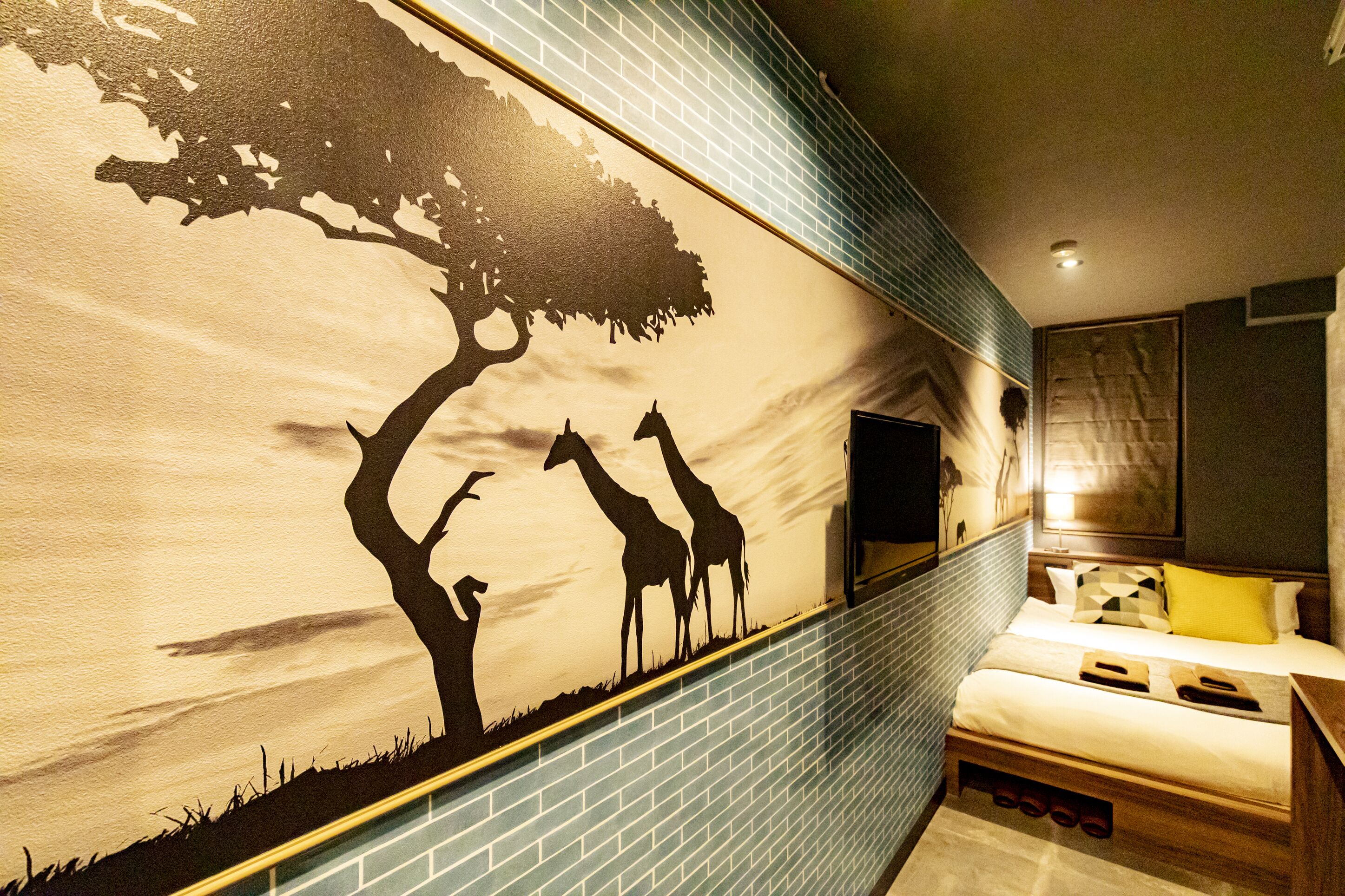 Une chambre d’hôtel ornée d’une fresque représentant des girafes et un arbre, un lit avec une tête de lit en bois, et un téléviseur à écran plat fixé au mur.