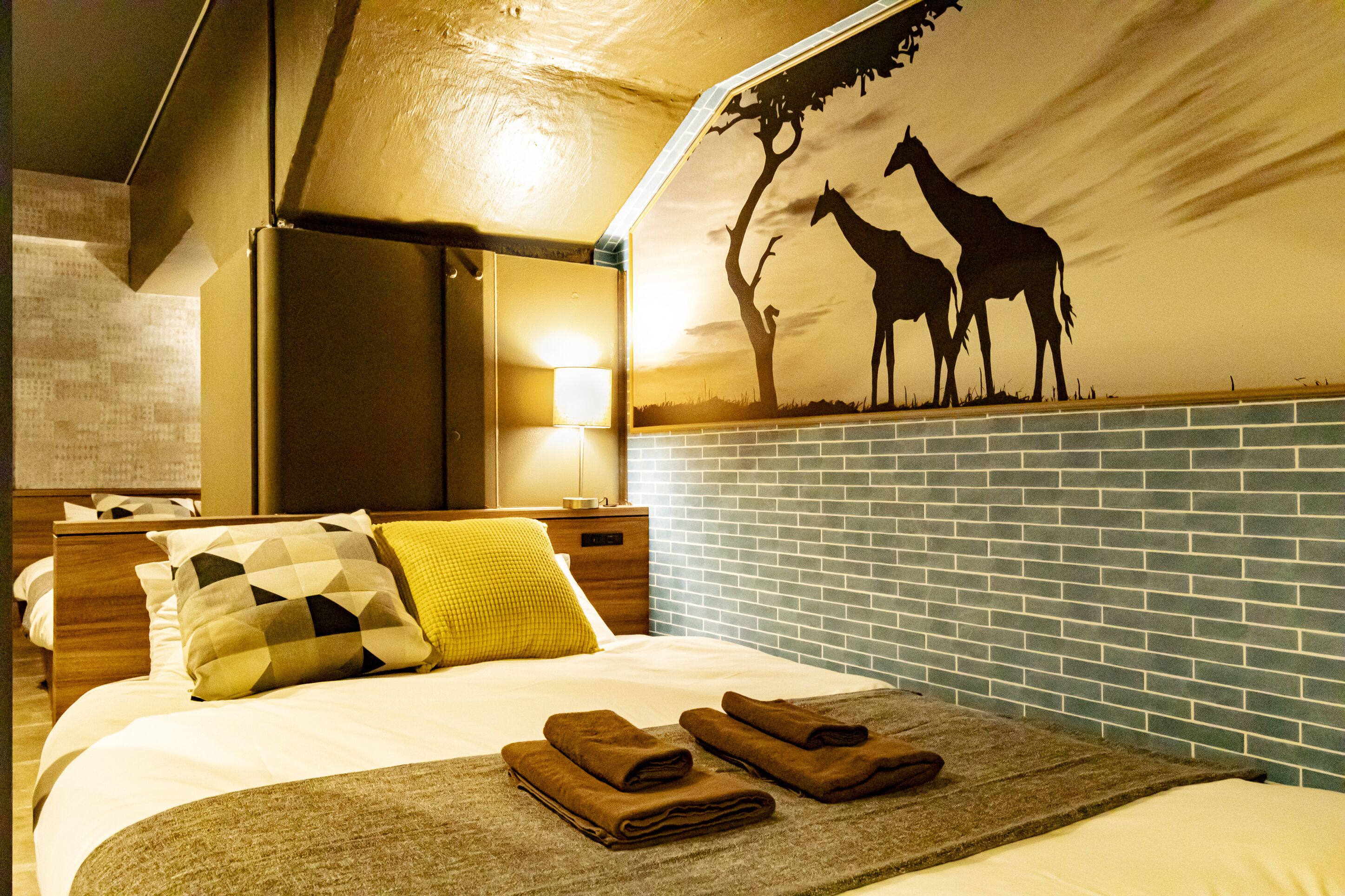 A hotel room with a bed, a nightstand, a wall mural of giraffes, and a blue-tiled wall.