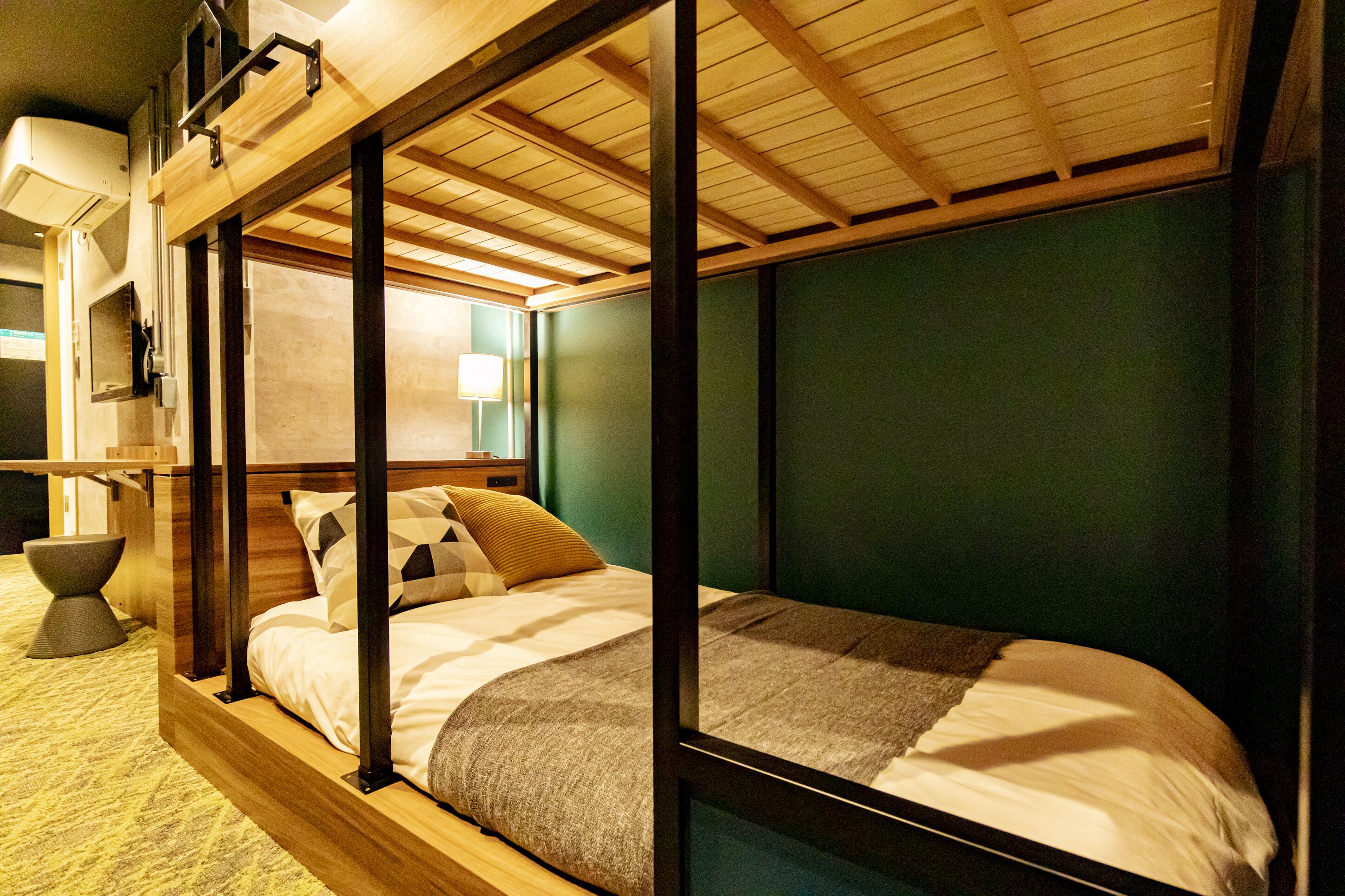 Cama litera con estructura de madera y pared verde de fondo. Hay una mesita de noche con una lámpara y una silla.