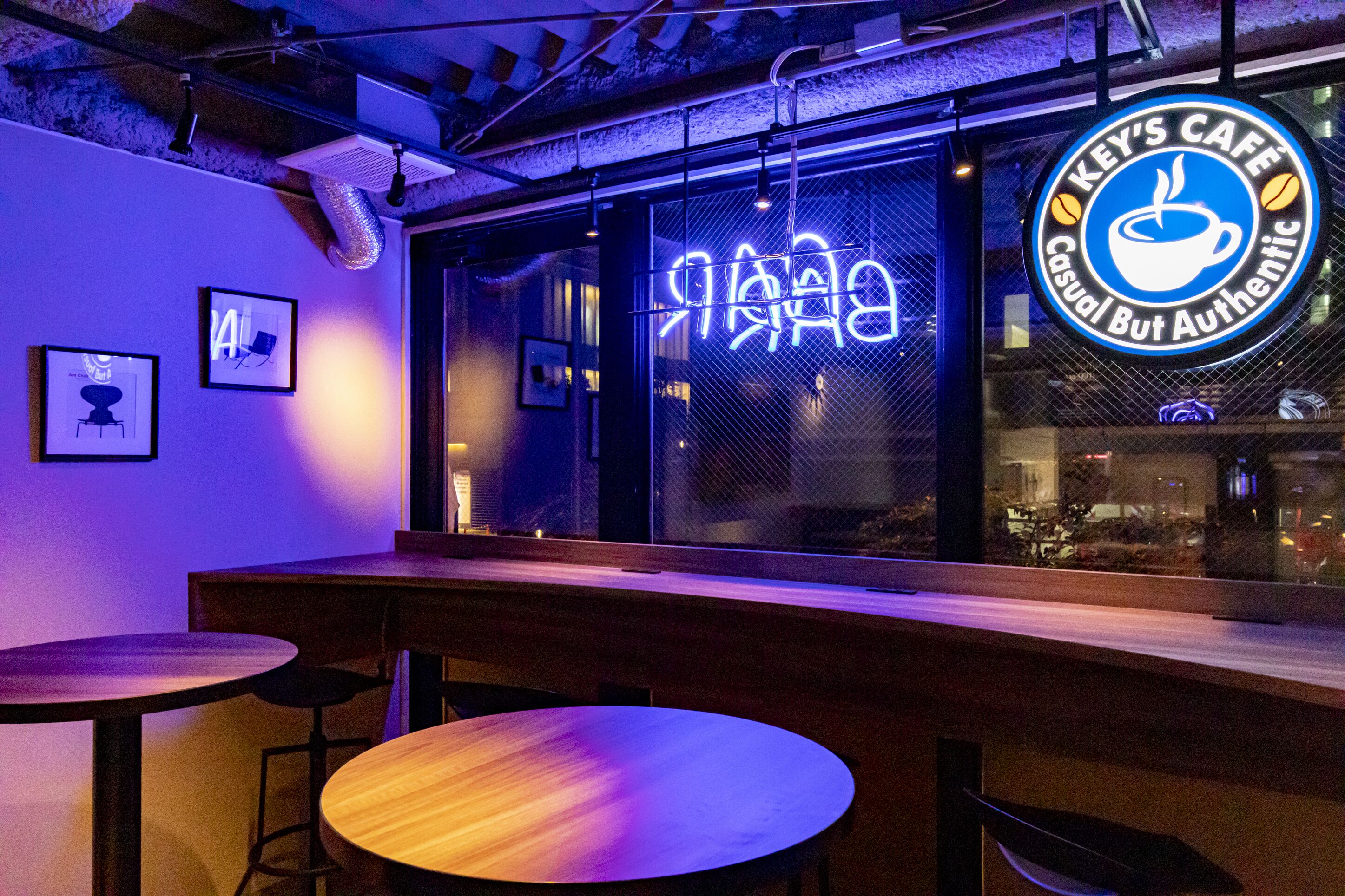 A cafe interior with tables and chairs, a neon sign reading "Key's Cafe Casual But Authentic," and a window with a view of the street outside.