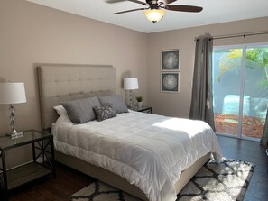 Een slaapkamer met een groot bed, twee nachtkastjes, een plafondventilator en uitzicht op een palmboom door een schuifdeur van glas.