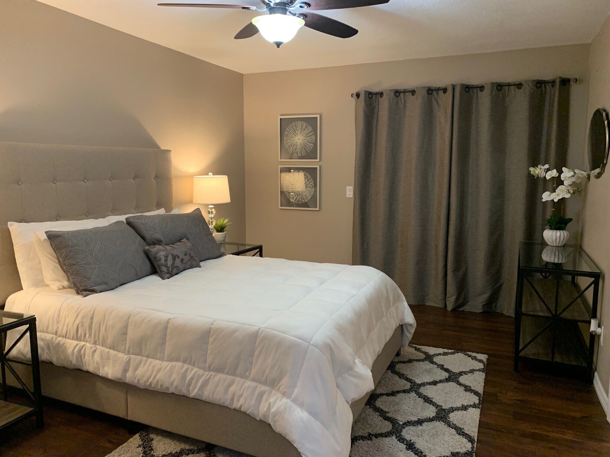 A bedroom with a bed, a ceiling fan, a nightstand, a lamp, a wall clock, and a window with curtains.