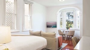 A bedroom with a large bed, a sofa, and a small table with chairs. There is a window with a view of trees and a painting on the wall.