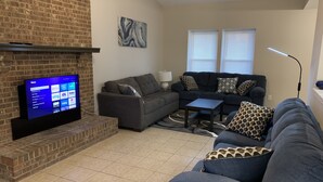 A living room with a brick fireplace, a flat-screen TV, a grey sofa, and patterned cushions. - Amazing home for 6 ! (Charlotte)