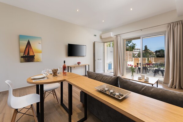A modern dining area with a wooden table, white chairs, and a sofa. A flat-screen TV is mounted on the wall, and a painting of a sailboat is visible.
