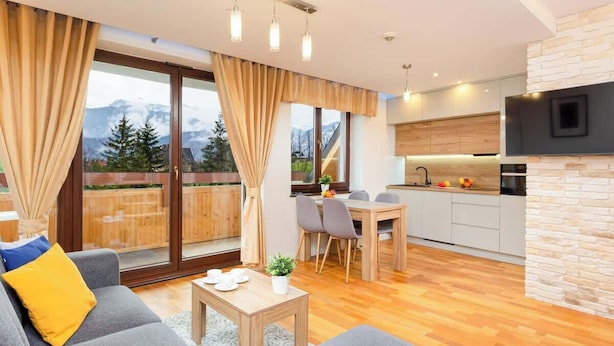 A modern living room with a sofa, a dining table, and a kitchen area with a stone wall and a flat-screen TV. - Suite with Mountain View | Gym | Sauna. (Koscielisko)