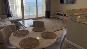 A modern living room with a sofa, a dining table with placemats, a TV mounted on the wall, and a balcony with a view of the sea.