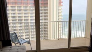 A room with a large sliding glass door leading to a balcony with a view of the ocean and high-rise buildings. There is a cushioned seating area and a glass coffee table.