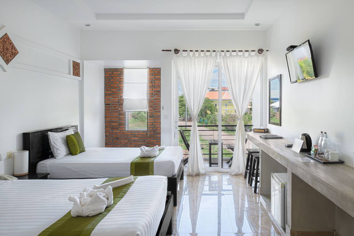 A hotel room with two beds, a balcony with a view, a desk with a coffee machine, and a TV mounted on the wall.