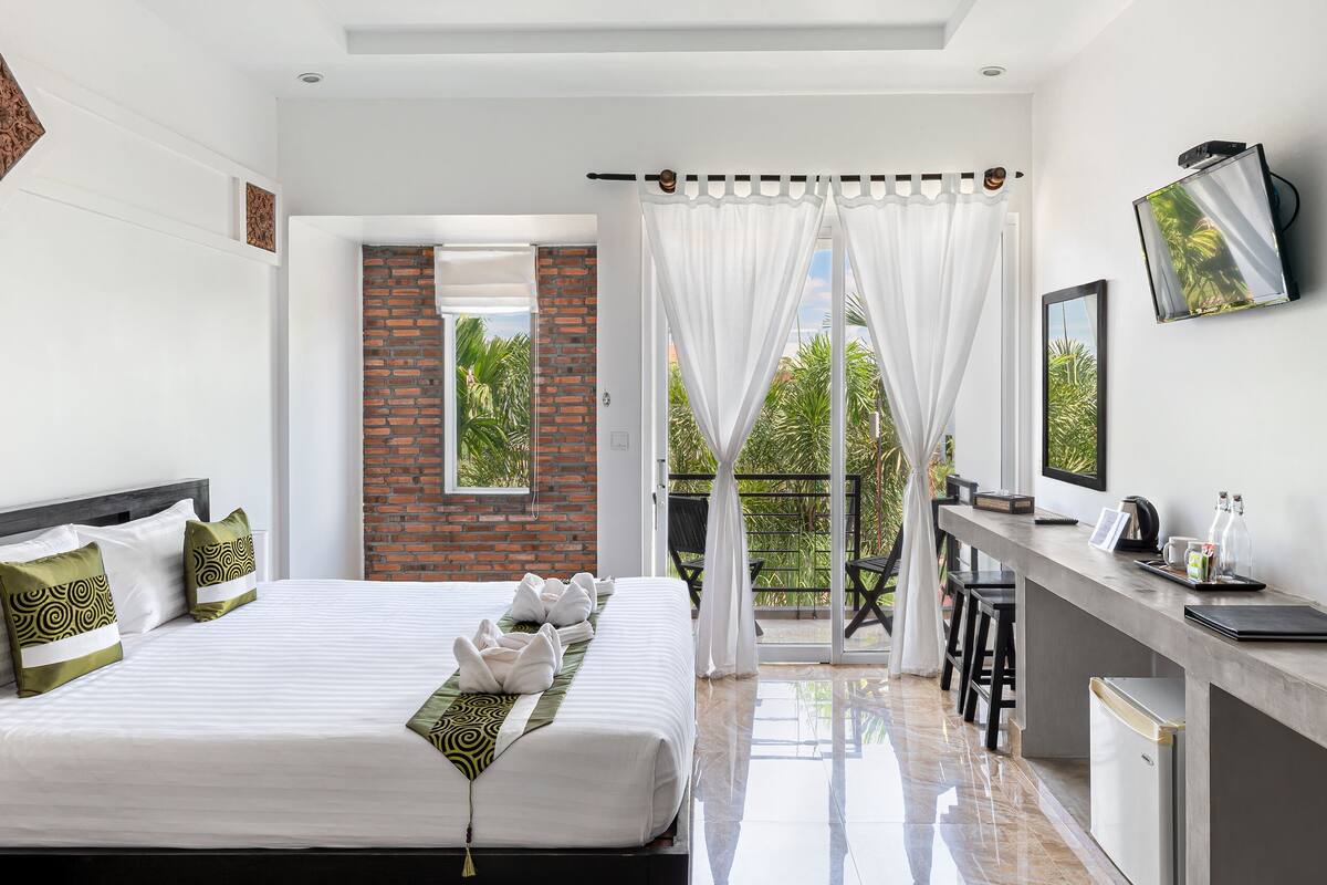 A modern hotel room with a large bed, a desk with a TV, and a balcony with a view of greenery.