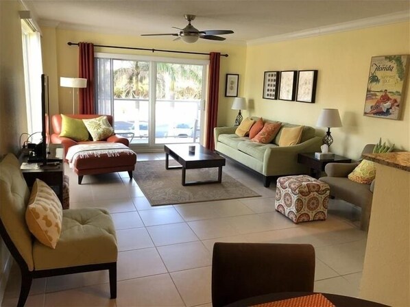 A living room with a sofa, armchair, coffee table, and a patterned ottoman. There is a ceiling fan, a window with curtains, and framed pictures on the wall. - Beachcomber unit 207 (Longboat Key)