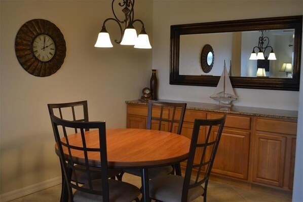 A dining area with a round wooden table, four chairs, a wall clock, and a mirror above the counter. - Beachcomber unit 605 (Longboat Key)