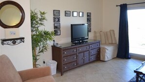 A living room with a television on a wooden cabinet, a potted plant, a sofa, and a window with curtains. - Beachcomber unit 605 (Longboat Key)