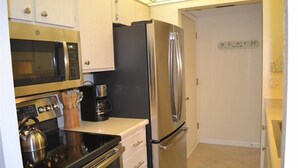 A compact kitchen with stainless steel appliances, including a microwave, oven, and refrigerator, with a sign reading "BEACH GEAR" on the wall. - Beachcomber unit 605 (Longboat Key)