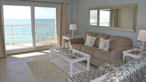 A living room with a large window overlooking the ocean, a beige sofa, a patterned ottoman, and a white coffee table. - Beachcomber unit 604 (Longboat Key)