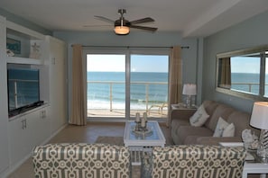A living room with a large window overlooking the ocean, a ceiling fan, a television, and a patterned sofa.