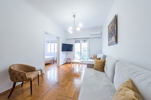 A modern living room with a white sofa, a brown armchair, a glass coffee table, and a flat-screen TV mounted on the wall.