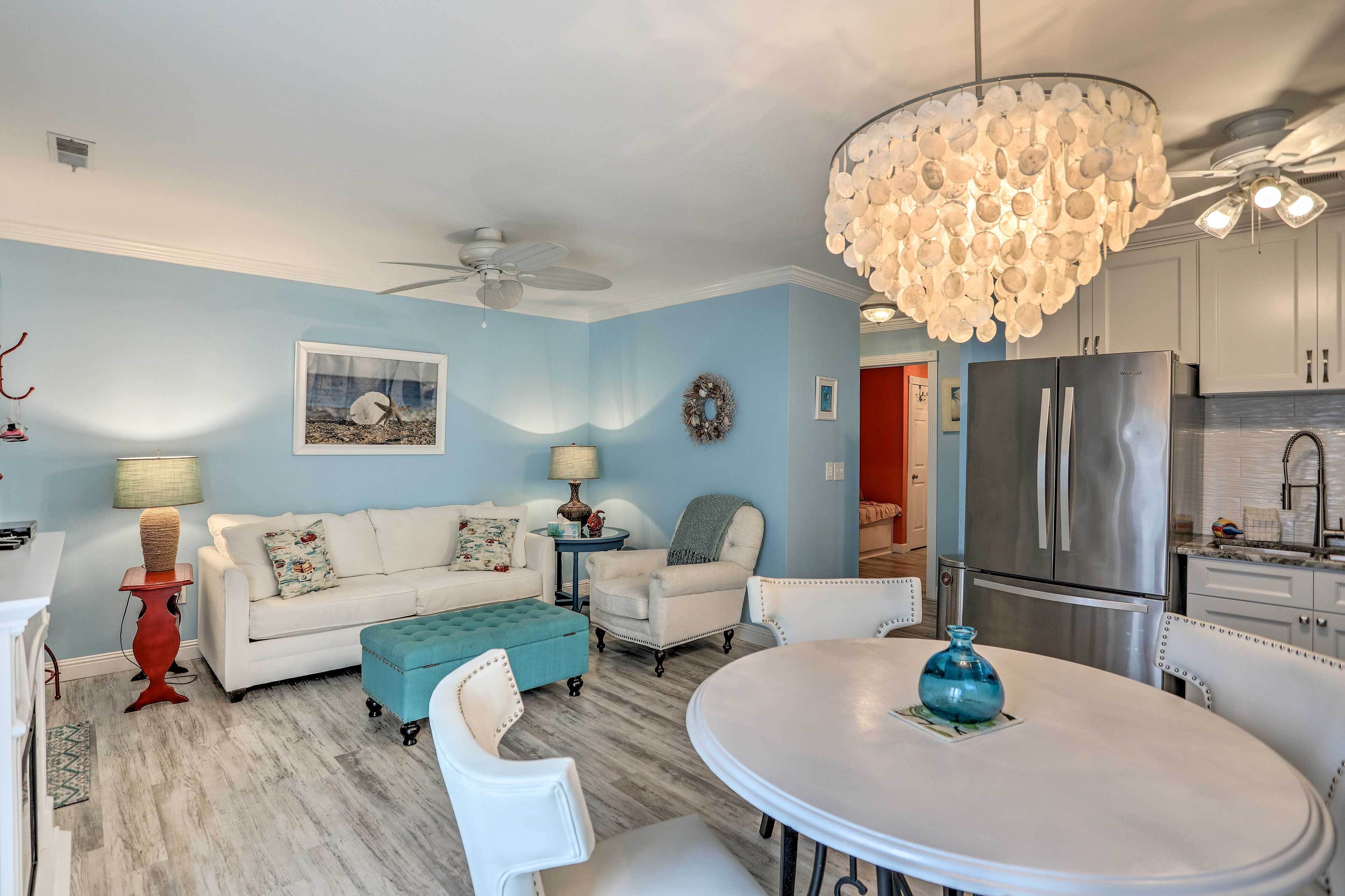 A living room with a white sofa, a teal ottoman, a round table with a blue vase, and a chandelier.