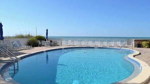 A clear blue swimming pool with a curved edge, surrounded by lounge chairs and umbrellas, with a view of the ocean in the background. - Beachcomber unit 502 (Longboat Key)