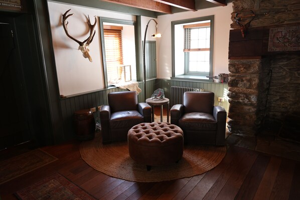 Une pièce chaleureuse avec des fauteuils en cuir, un pouf rond, une cheminée et un crâne de cerf accroché au mur.