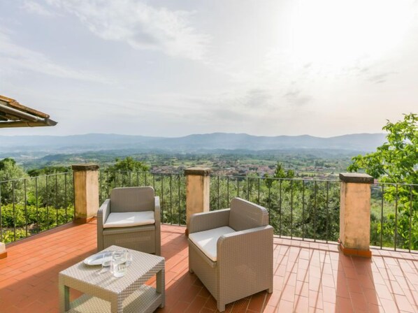 Une terrasse dotée de meubles en osier et d’une table, offrant une vue panoramique sur un paysage pittoresque avec des collines et une végétation luxuriante.