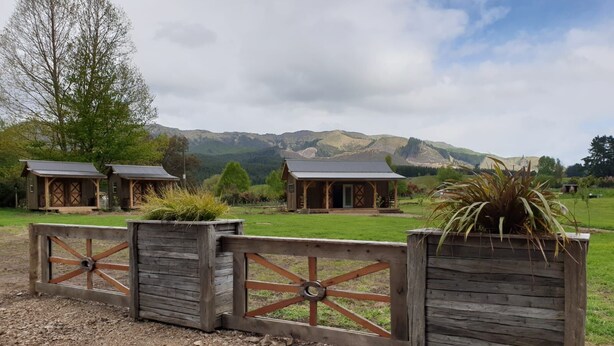 A rustic wooden fence with a gate, leading to a property with multiple cabins in a mountainous landscape. - Mohaka River Farm - Campsite (Te Haroto)