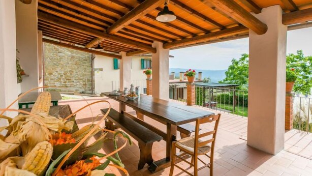A covered outdoor dining area with a wooden table and chairs, a stone wall, and a view of the sea. - Appartamento Piccolo by Interhome (Reggello)
