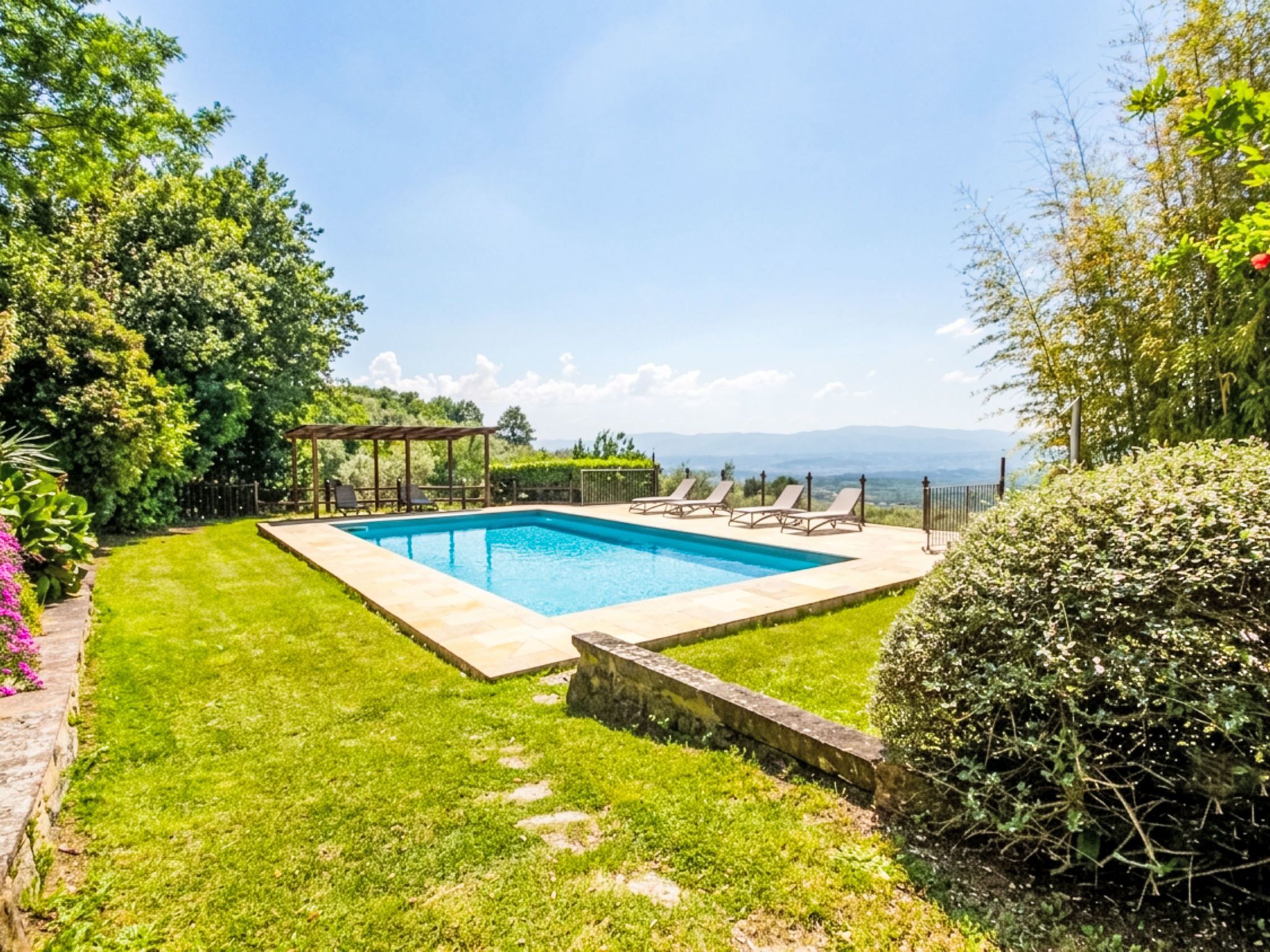 Una piscina rectangular con un pérgola y sillas de descanso, rodeada por un césped cuidado y setos.