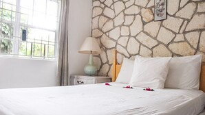A neatly made bed with white linens and rose petals, a stone wall, a lamp, and a small nightstand with a decorative item. - View 61 Hibiscus with Private pool & Beach View (St John's)