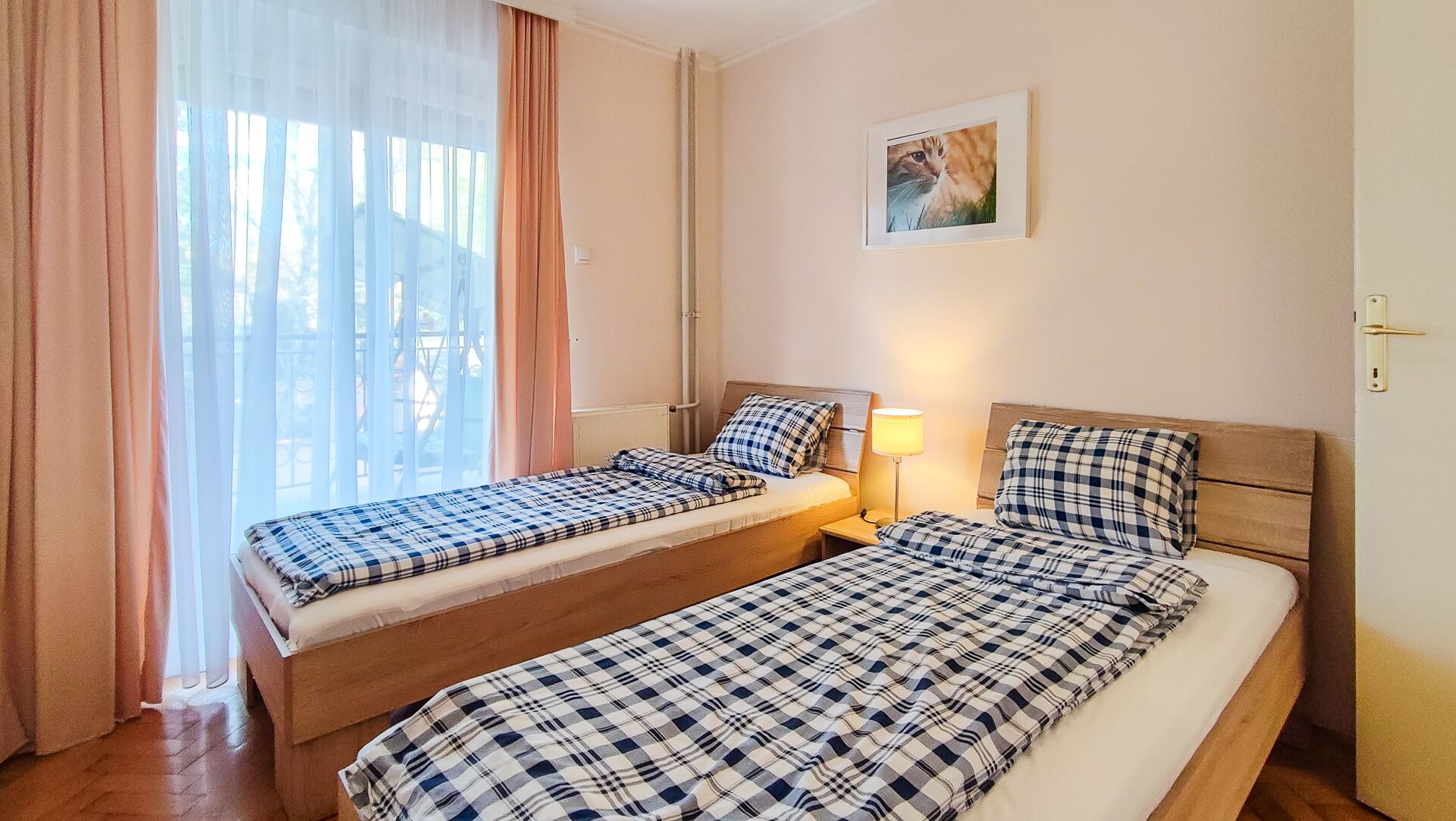 Une chambre avec deux lits, chacun recouvert d’une couvre-lit à carreaux, une table de chevet avec une lampe et une fenêtre ornée de rideaux légers.