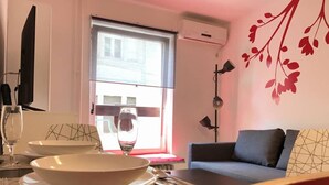 Una sala de estar moderna con comedor, un sofá, una mesa de comedor dispuesta con platos y cubiertos, una copa de vino y una ventana con vista a edificios.