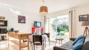 A modern living room with a sofa, a dining table, and chairs. There is a television on a stand, a wicker lamp, and a view of the garden through large windows. - Astrida by Interhome (Carnac)