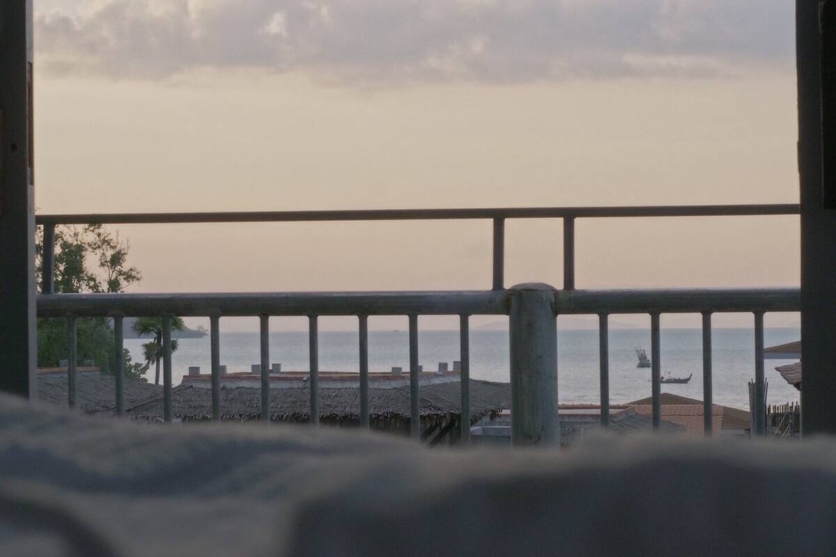 Un balcón con baranda metálica que da a una playa, con edificios y un barco a lo lejos.