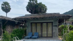 Une piscine avec une terrasse en bois, deux chaises longues et un petit bâtiment doté d’un toit de chaume.