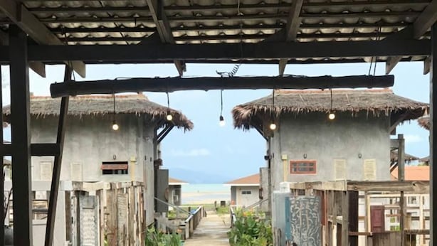 Une allée couverte mène à des bâtiments de style traditionnel, dotés de toits de chaume, et est entourée de verdure, offrant une vue dégagée sur la mer.