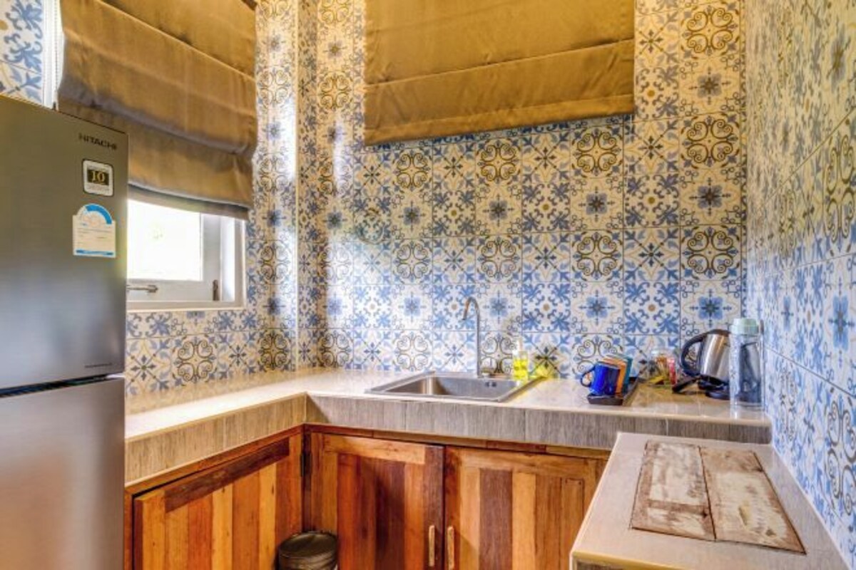 A kitchen with patterned blue and white tiles, a stainless steel refrigerator, wooden cabinets, and a window with a beige curtain.