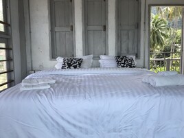 Una cama con sábanas blancas y dos almohadas con estampado blanco y negro, una ventana con vista a palmeras y una puerta de madera.