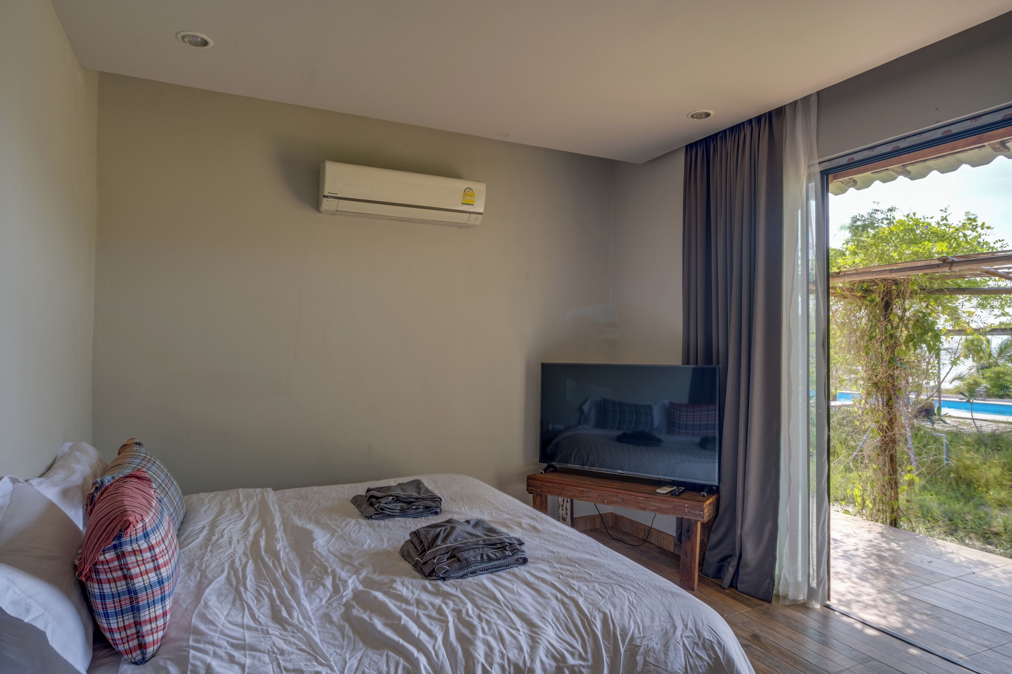 A bedroom with a bed, a TV on a wooden stand, an air conditioner on the wall, and a view of greenery outside.