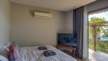 A bedroom with a bed, a TV on a wooden stand, an air conditioner on the wall, and a view of greenery outside.