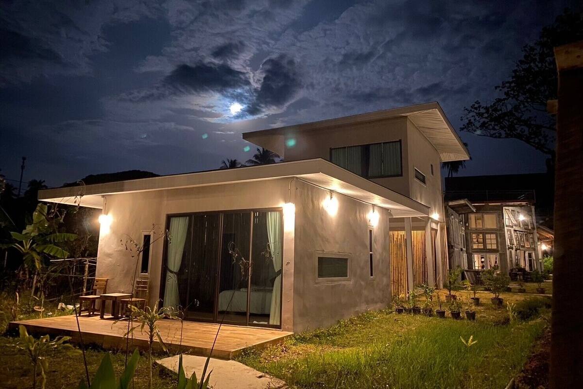 Una casa moderna de dos pisos con un porche cubierto y un dormitorio visible a través de puertas de vidrio.