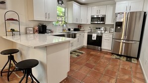 A modern kitchen with white cabinets, a stainless steel refrigerator, a microwave, and a sink. There are two black stools and a coffee machine on the counter.