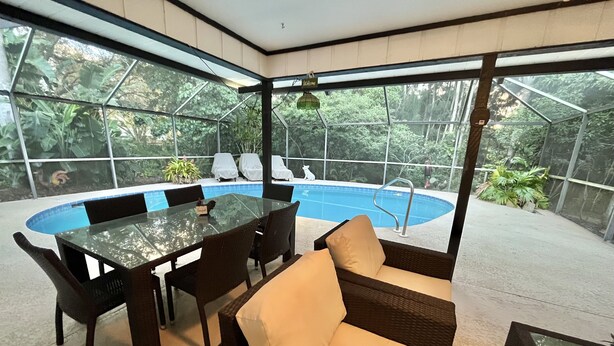 A modern outdoor dining area with a glass table and chairs, a pool, and a view of lush greenery.
