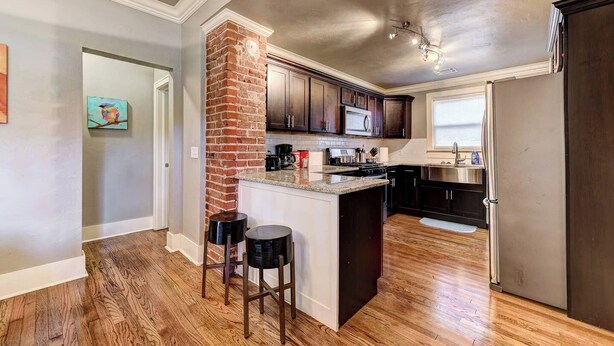 Una cocina con piso de madera, pared de ladrillo decorativa, gabinetes oscuros, refrigerador de acero inoxidable y ventana con persianas.