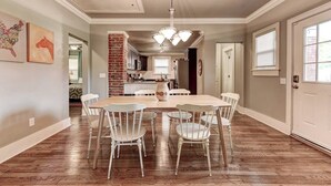 Un comedor con una mesa de madera y sillas blancas, una pared de ladrillo decorativa y una cocina al fondo.