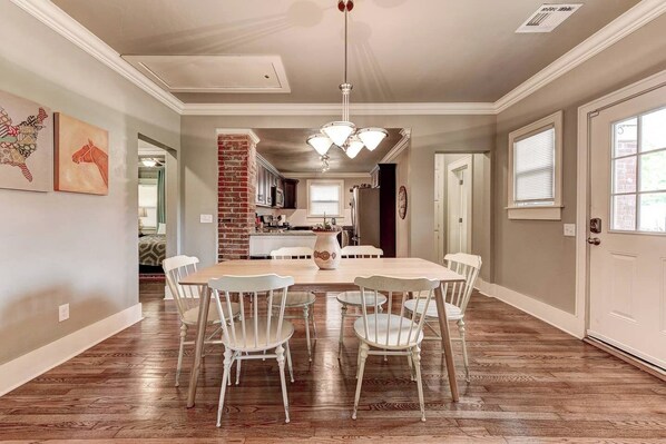 A dining room with a wooden table and white chairs, a brick accent wall, and a kitchen in the background.