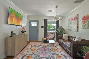 A living room with a sofa, a coffee table, a TV mounted on the wall, a door, and a window with curtains. - Communal Crashpad (Oklahoma City)