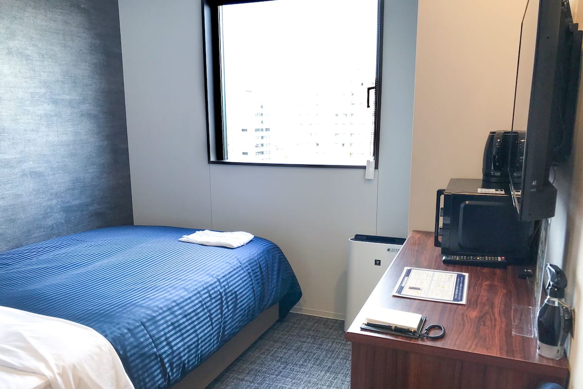 A hotel room with a bed, a desk with a computer, a printer, and a window with a city view.