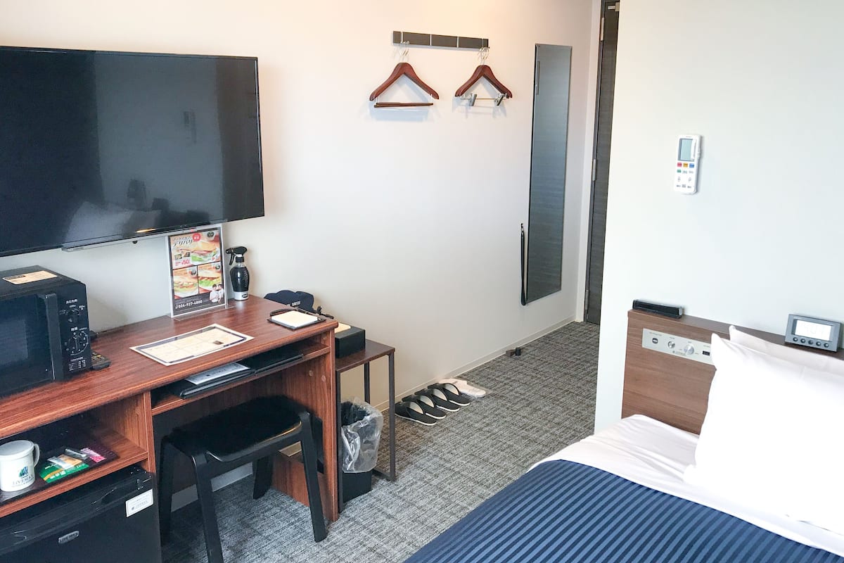 A hotel room with a bed, desk, chair, TV, coat hooks, and a shoe rack.